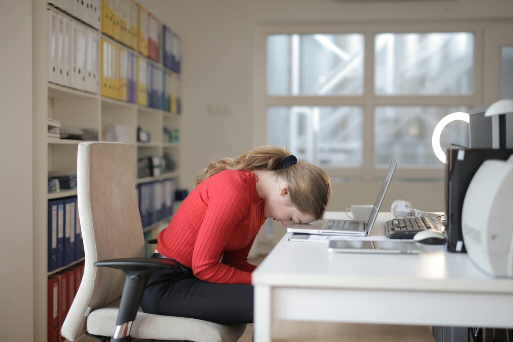 Tired woman in red sweater naps on office desk beside laptop, overwhelmed by remote work.